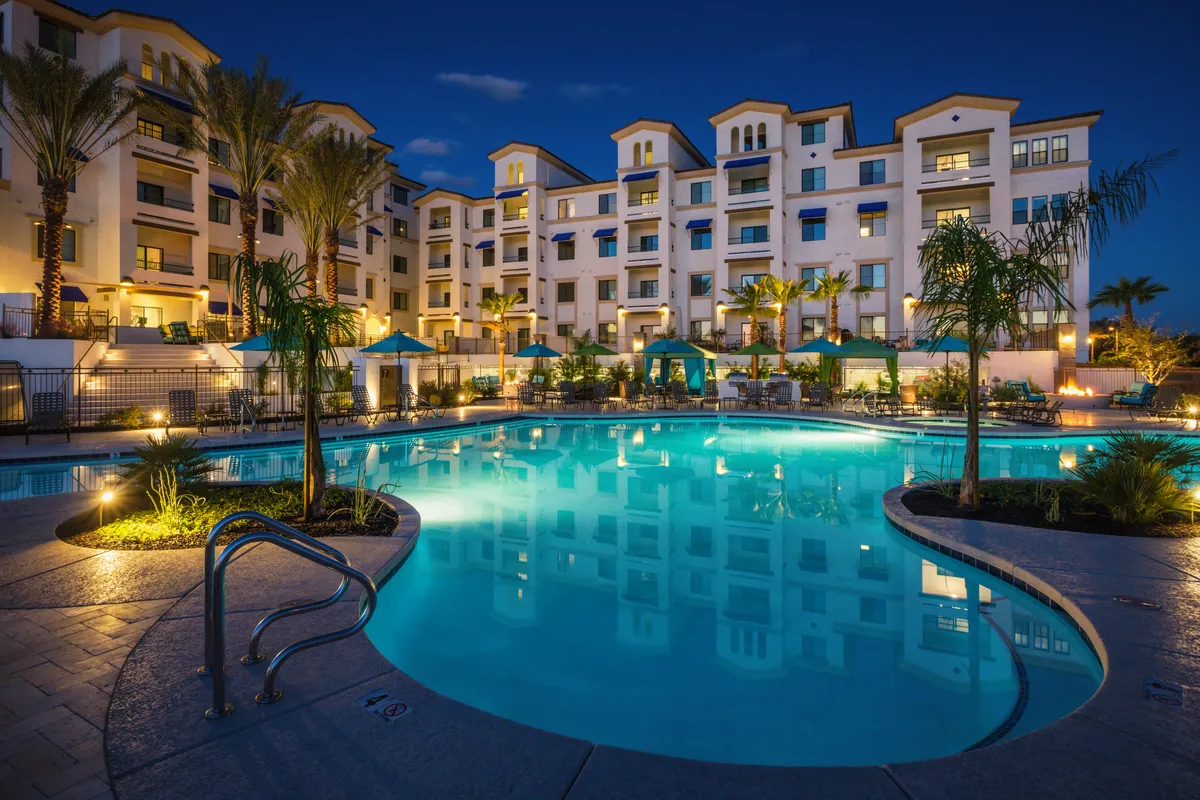 A luxurious outdoor swimming pool area surrounded by palm trees and lit umbrellas, with the building facade visible in the background. The scene is set at twilight, creating a warm and inviting ambiance for evening gatherings or relaxed stargazing by the water.