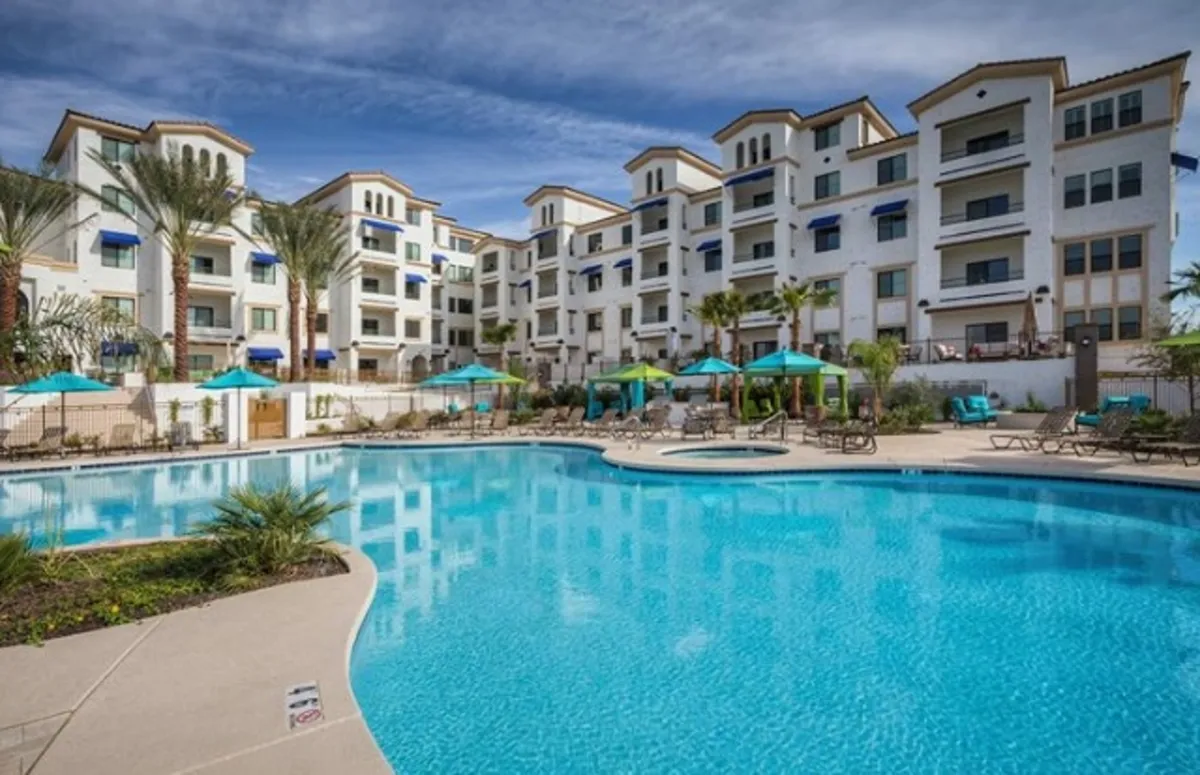 A relaxed poolside exterior with a large swimming pool, building facade, tall palm trees, and plenty of lounge chairs and umbrellas. The setting is ideal for swimming, sunbathing, or social gatherings, especially at sunset.