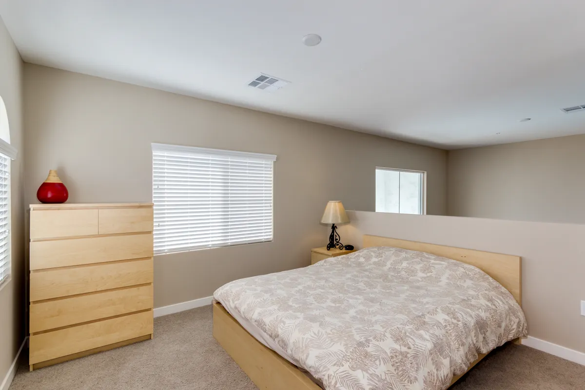 A tranquil bedroom scene with a bed dressed in patterned bedding, a wooden dresser, and a nightstand with a lamp. Natural light is visible, adding warmth and a sense of morning tranquility to the space.