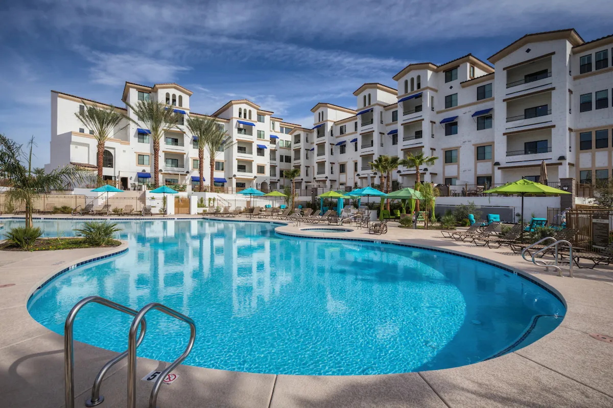 A sunlit exterior view of a swimming pool surrounded by palm trees, comfortable lounge chairs, and the building's modern facade. The environment invites socializing, sunbathing, and relaxed gatherings by the water.