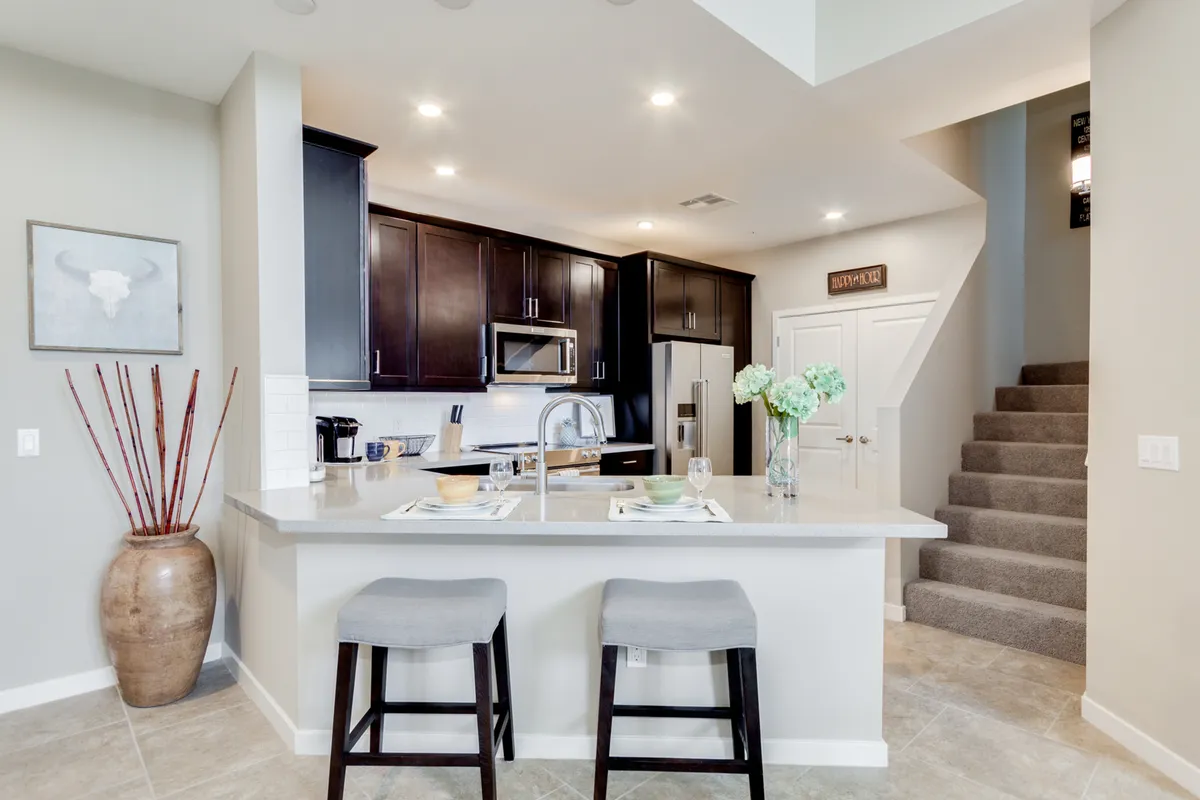 A bright, inviting kitchen scene with a spacious island, stools for casual dining, visible kitchen appliances, a decorative vase adding a homey touch, and an open view toward a staircase. The open layout encourages socializing and shared meals.