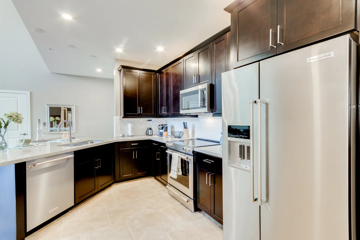 This image features a contemporary kitchen with stainless steel appliances, dark cabinetry, and a sleek countertop with an integrated sink. The space is warmly lit, showcasing high-end finishes and a practical layout ideal for cooking and entertaining.