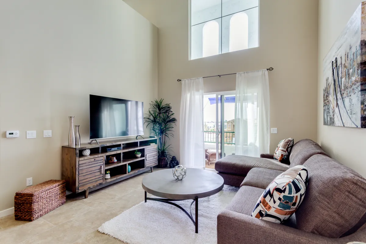 A cozy living room with a plush sofa, coffee table, TV, and curtains framing a balcony view. The scene is bathed in natural light, highlighting the inviting space for casual get-togethers and quiet relaxation, with a hint of outdoor views through the windows.