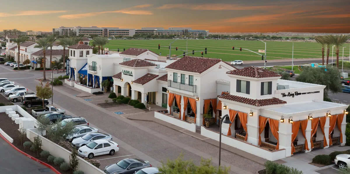A vibrant exterior scene featuring colorful awnings, palm trees, well-maintained walkways, and a spacious parking area. People can be seen dining outdoors, socializing, and browsing nearby boutiques, embodying the energy and attractions of the neighborhood.
