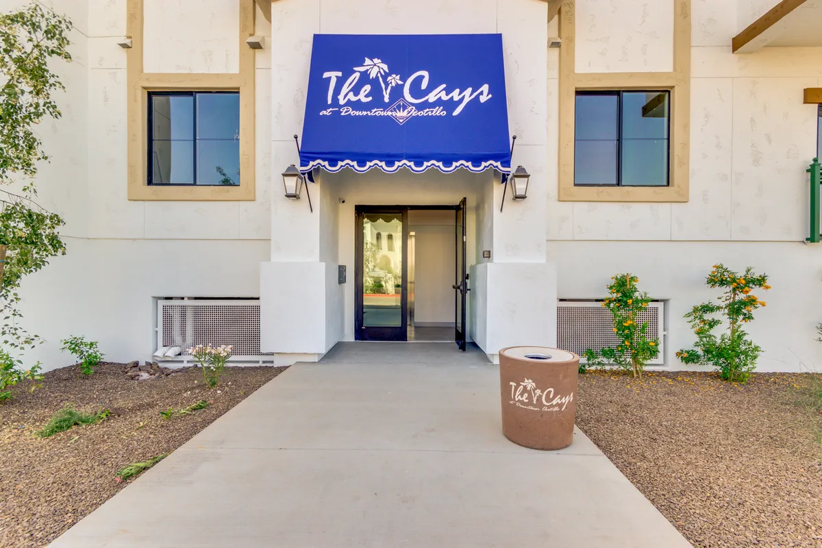 A welcoming exterior view of the property's entrance, featuring a canopy awning, visible signage, beautifully landscaped greenery, and a broad doorway. The setting exudes a sense of arrival, safety, and community.