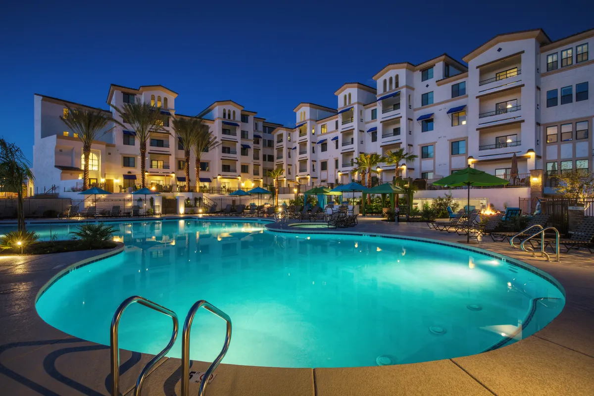 An expansive exterior shot showcasing the glistening swimming pool bordered by palm trees and outdoor seating, all set against the contemporary building facade. The area is designed for both relaxation and socializing, ideal for evening gatherings or starlit swims.
