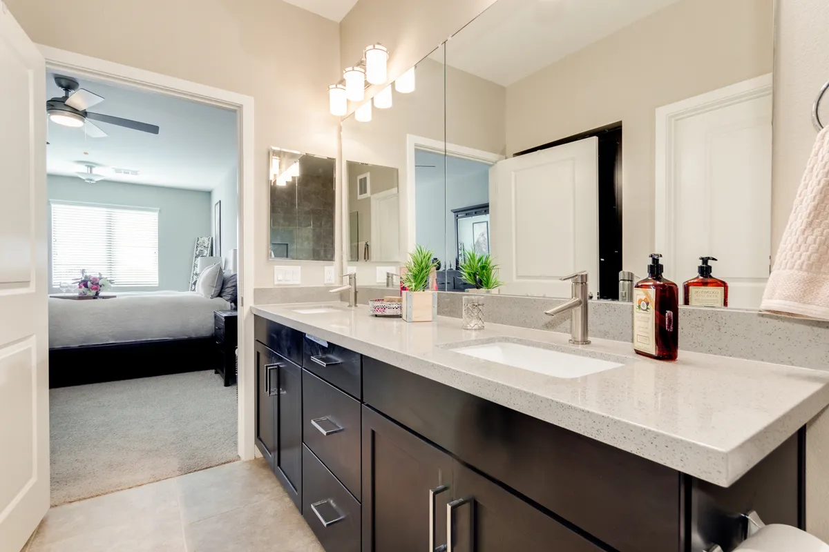 A spacious bathroom scene featuring a double sink vanity, a large mirror, decorative plants, and bright lighting. The atmosphere is fresh, functional, and stylish.