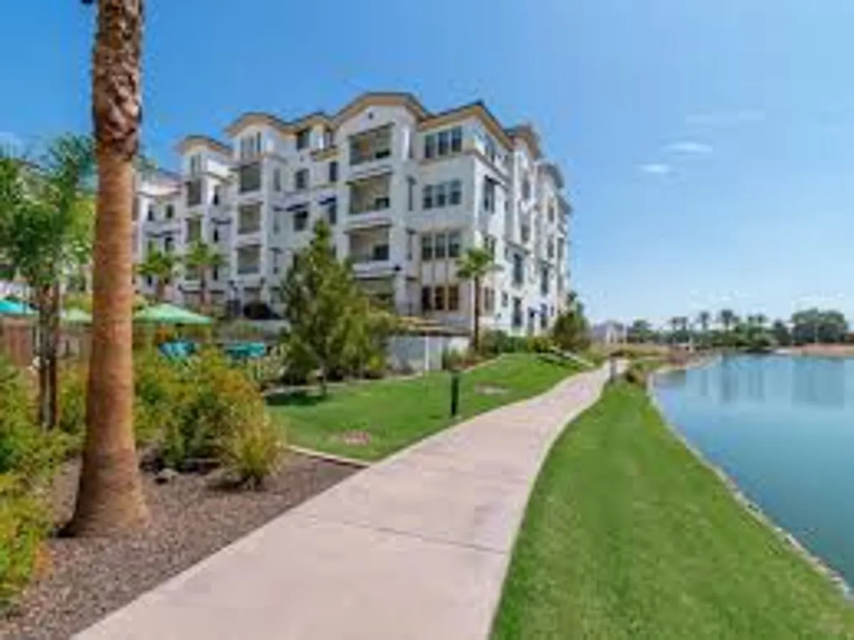 A tranquil exterior shot of a modern apartment building surrounded by lush greenery, with a pathway running alongside a serene water feature. The setting sun reflects on the water, creating a peaceful neighborhood ambiance ideal for evening walks and outdoor relaxation.